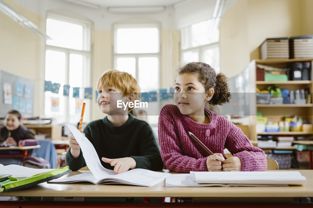 Smiling blond boy sitting with girl at desk looking away in classroom