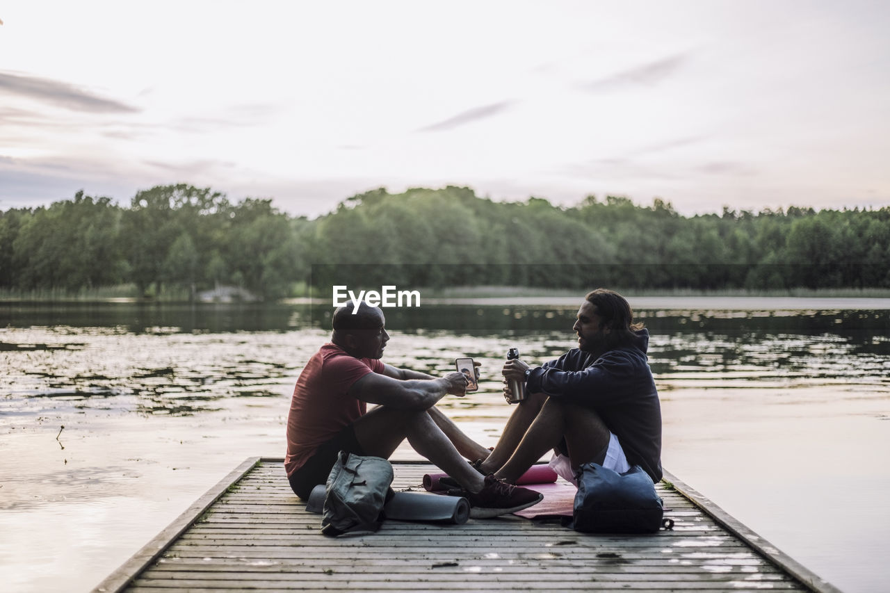 Man sharing smart phone with friend sitting on jetty at lake