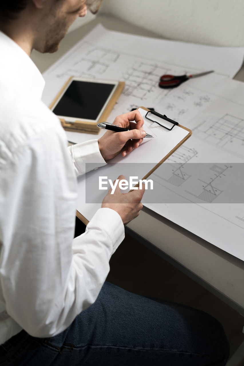 Male graphic designer writing in clipboard while sitting at desk in office