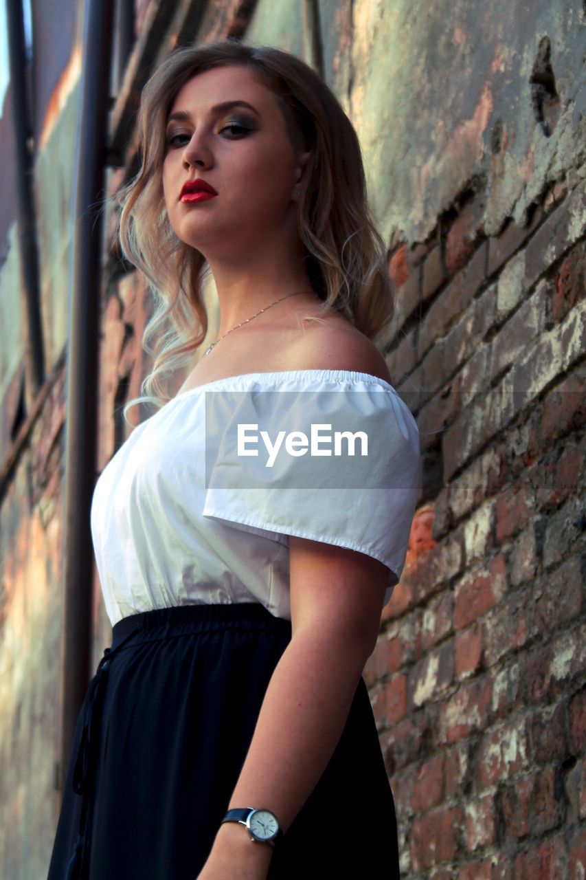 Low angle portrait of woman standing by weathered brick wall