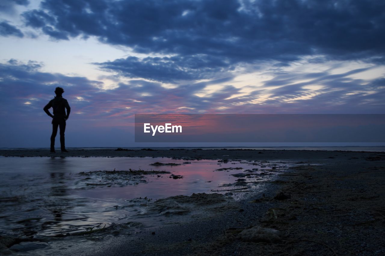 SILHOUETTE MAN STANDING ON BEACH AGAINST SKY AT SUNSET