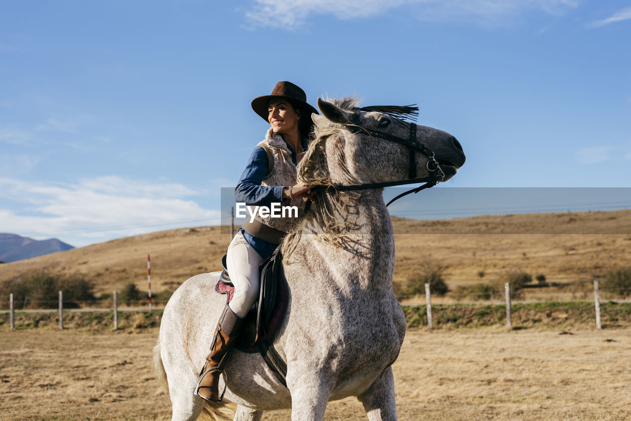 Side view of woman in hat sitting on beautiful white horse on background of rural landscape under blue sky