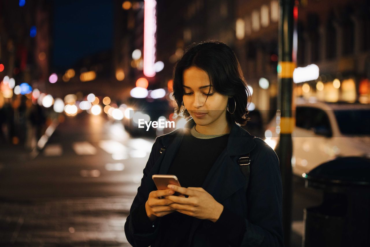 Young woman using mobile phone while standing in city at night