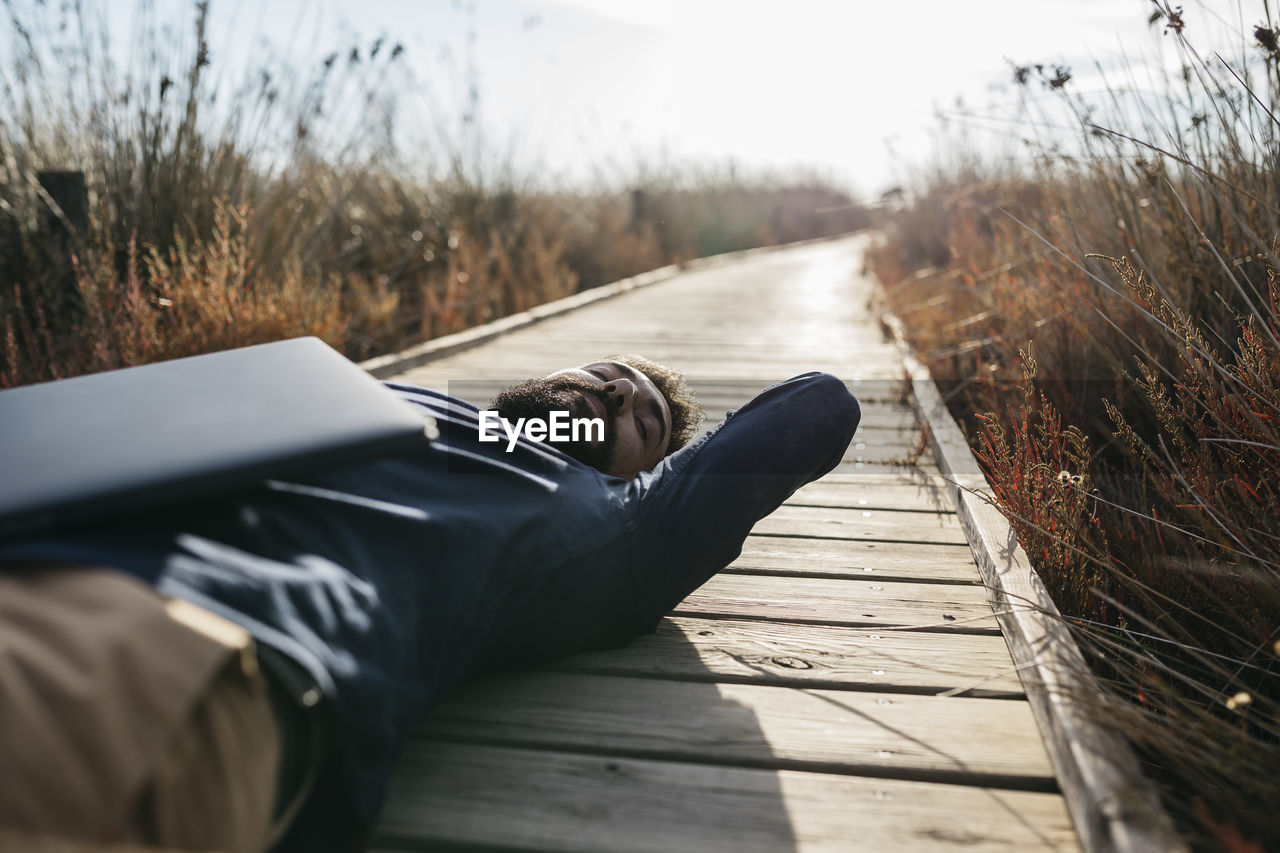 Man with laptop lying down amidst field on boardwalk during sunny day