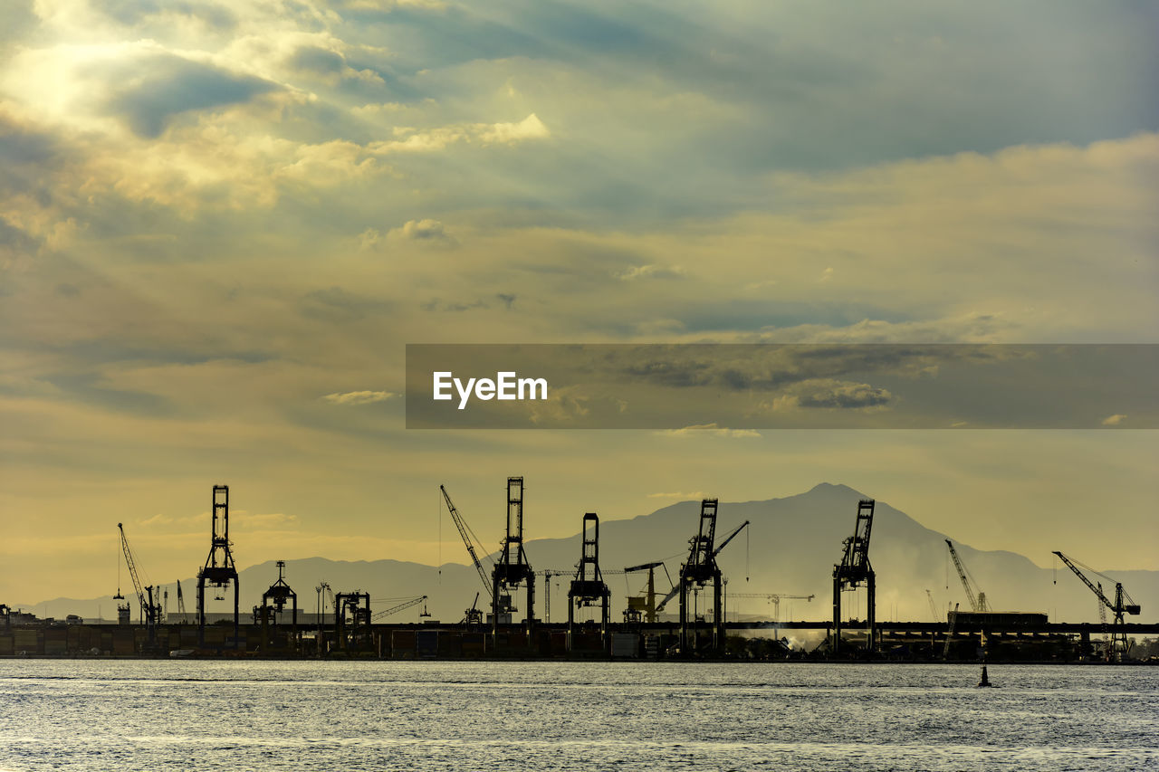 Silhouette cranes at harbor against sky during sunset