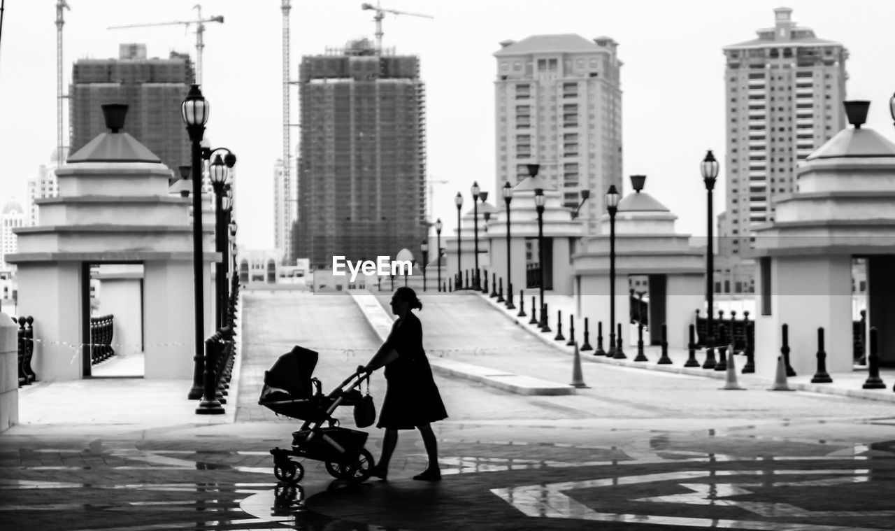 Side view of mother with baby carriage walking against buildings in city