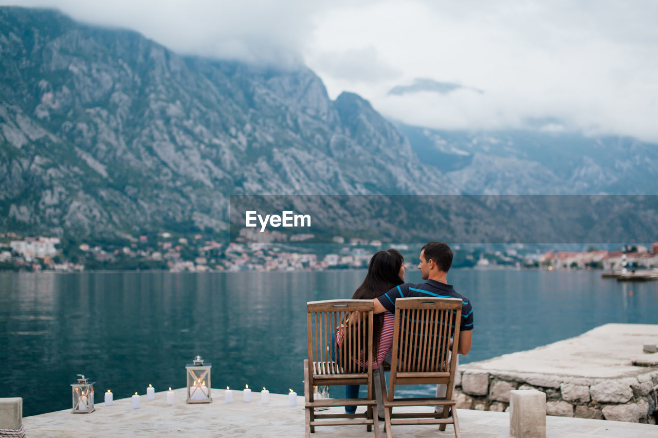 Rear view of couple sitting on lakeshore against mountains