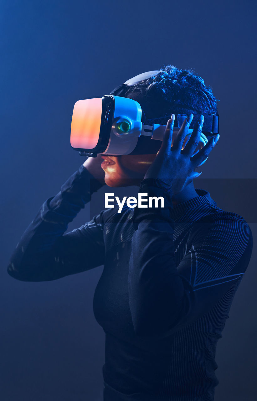 Astonished female african american gamer in modern vr goggles standing on dark background in studio while experiencing virtual reality in neon illumination