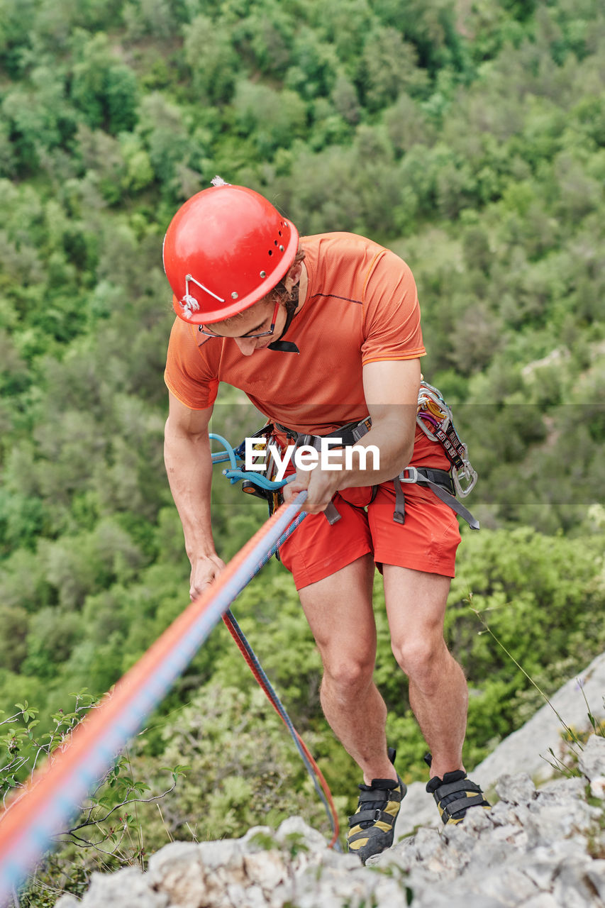 From above of brave male alpinist in protective equipment climbing steep cliff on background of forest