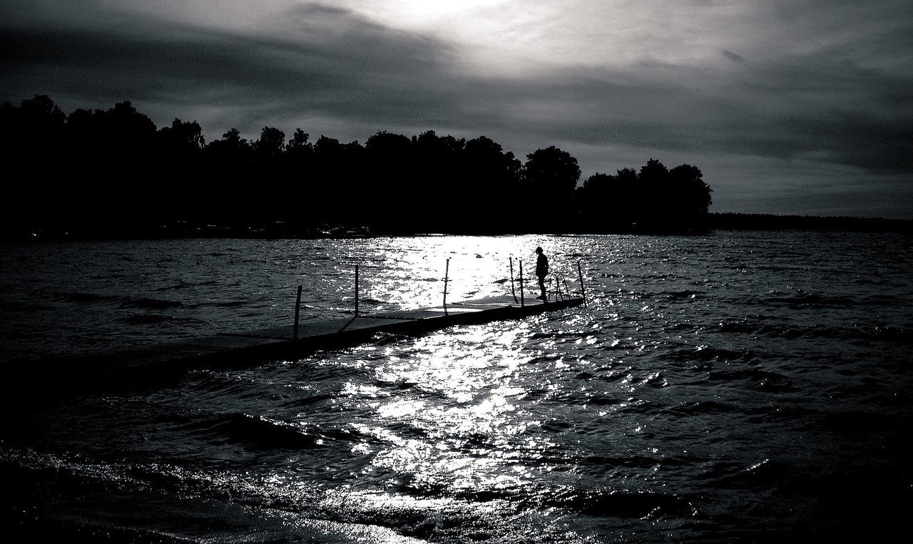 Silhouette woman standing on boardwalk at lake against cloudy sky