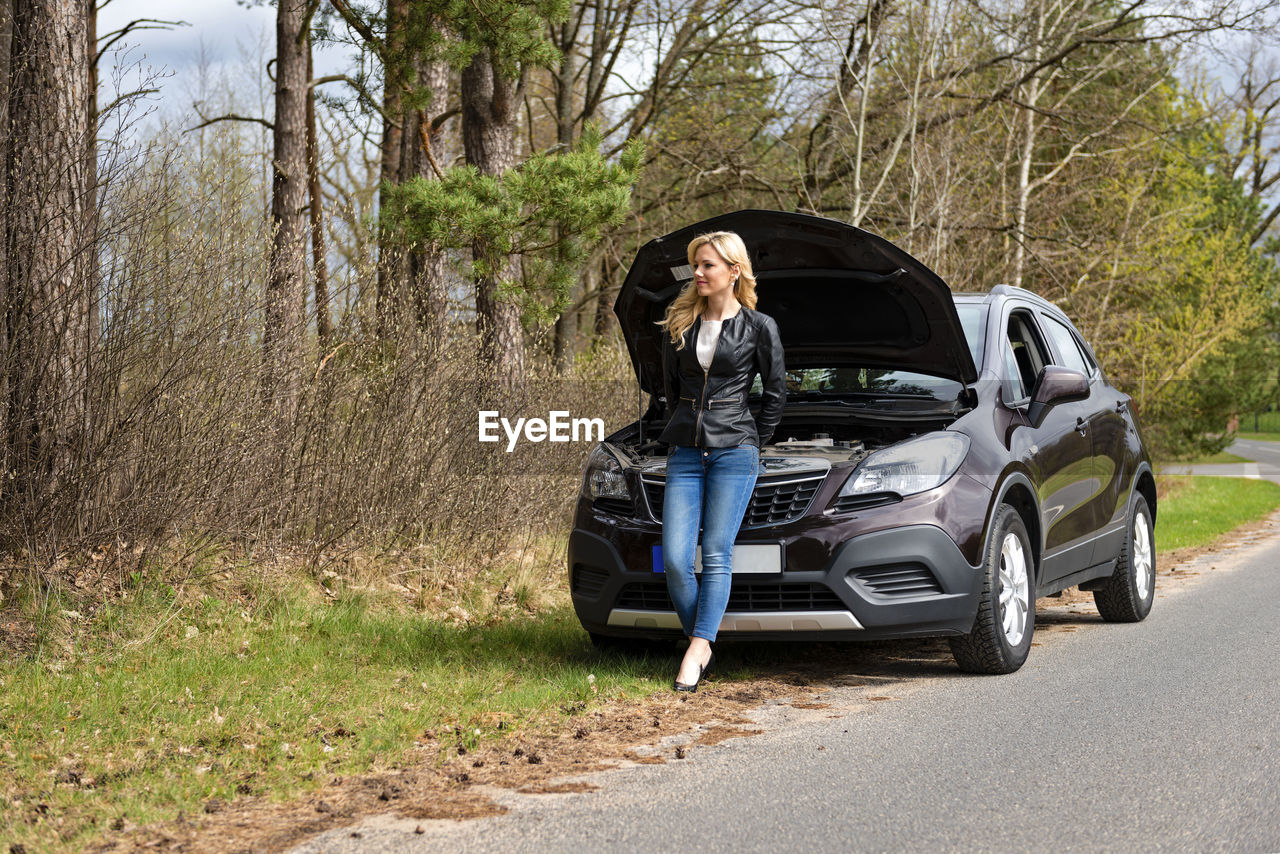 Full length of fashionable young woman standing by breakdown car on roadside