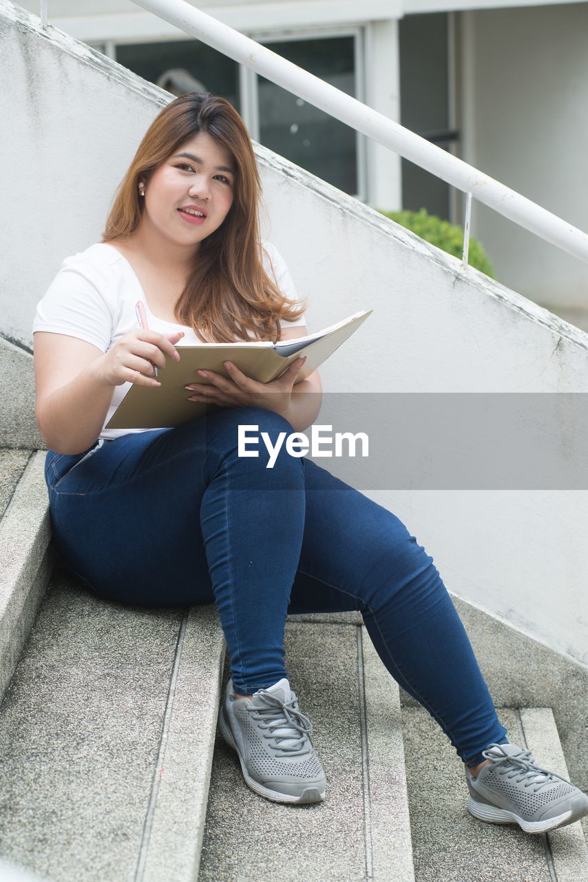 Full length portrait of young woman holding book while sitting on steps