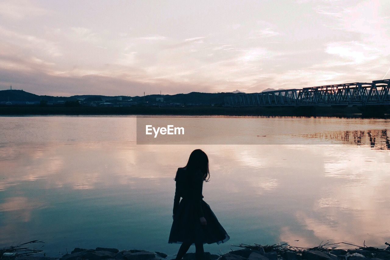 Silhouette girl standing by lake against sky during sunset