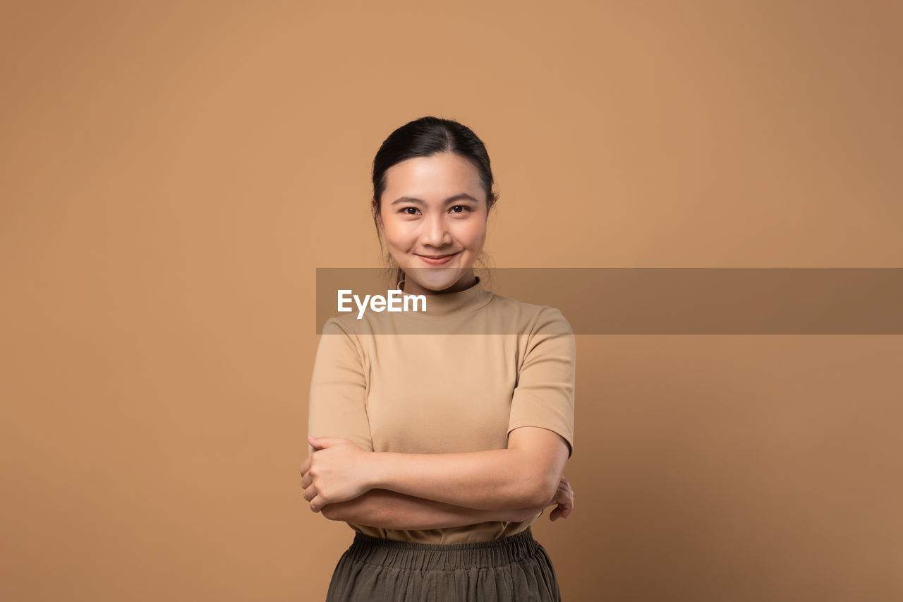 portrait, looking at camera, smiling, studio shot, one person, indoors, happiness, colored background, women, adult, emotion, arms crossed, standing, copy space, front view, young adult, waist up, cheerful, female, photo shoot, hairstyle, person, teeth, smile, positive emotion, casual clothing, three quarter length, individuality, brown hair, fashion, cut out, clothing