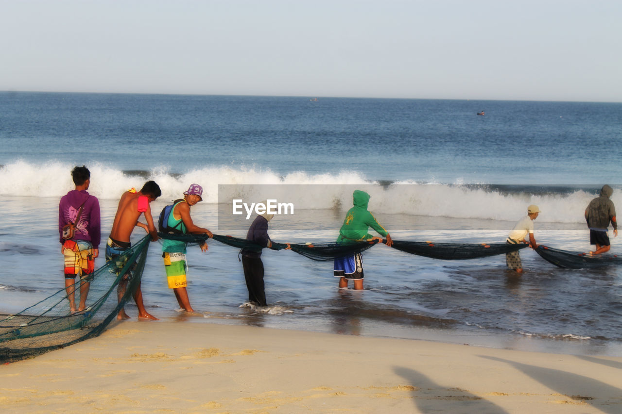 water, sea, group of people, beach, horizon over water, horizon, sky, nature, men, land, shore, ocean, coast, wave, nautical vessel, day, adult, vehicle, transportation, motion, boating, women, occupation, sand, group, beauty in nature, full length, scenics - nature, sports, teamwork, cooperation, outdoors, standing, boat, medium group of people, togetherness, fisherman, clear sky, body of water, paddle, water sports, lifestyles, leisure activity