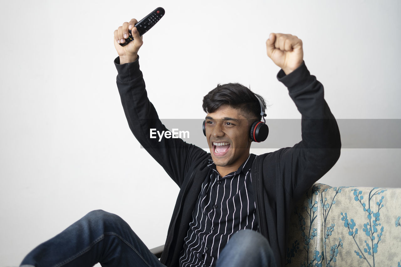 Asian man with headphone wearing black jacket holding remote while cheering for indian team.