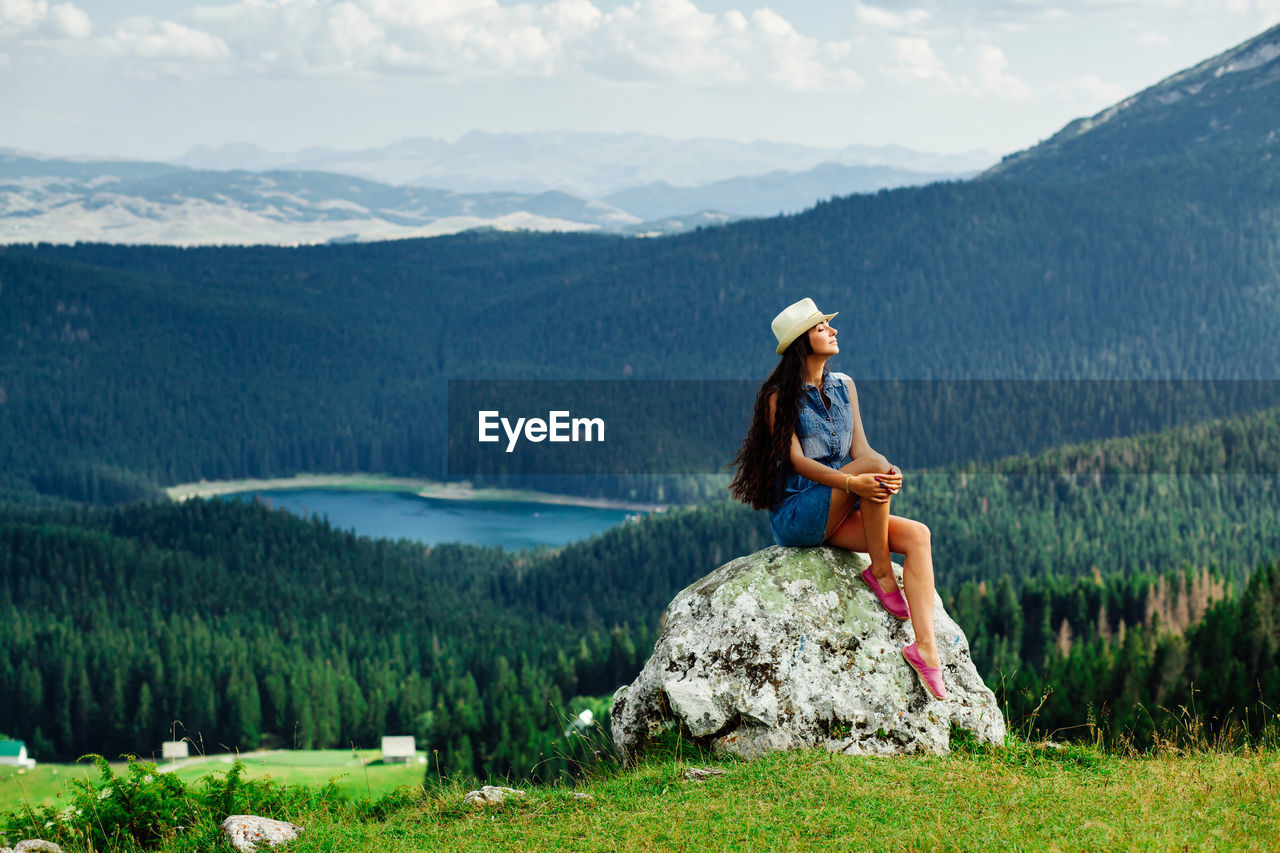 Full length of young woman sitting on rock against mountains