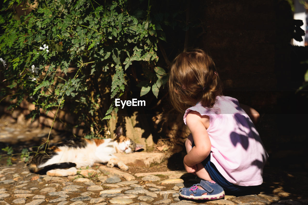 Rear view of girl crouching by cat sleeping in yard