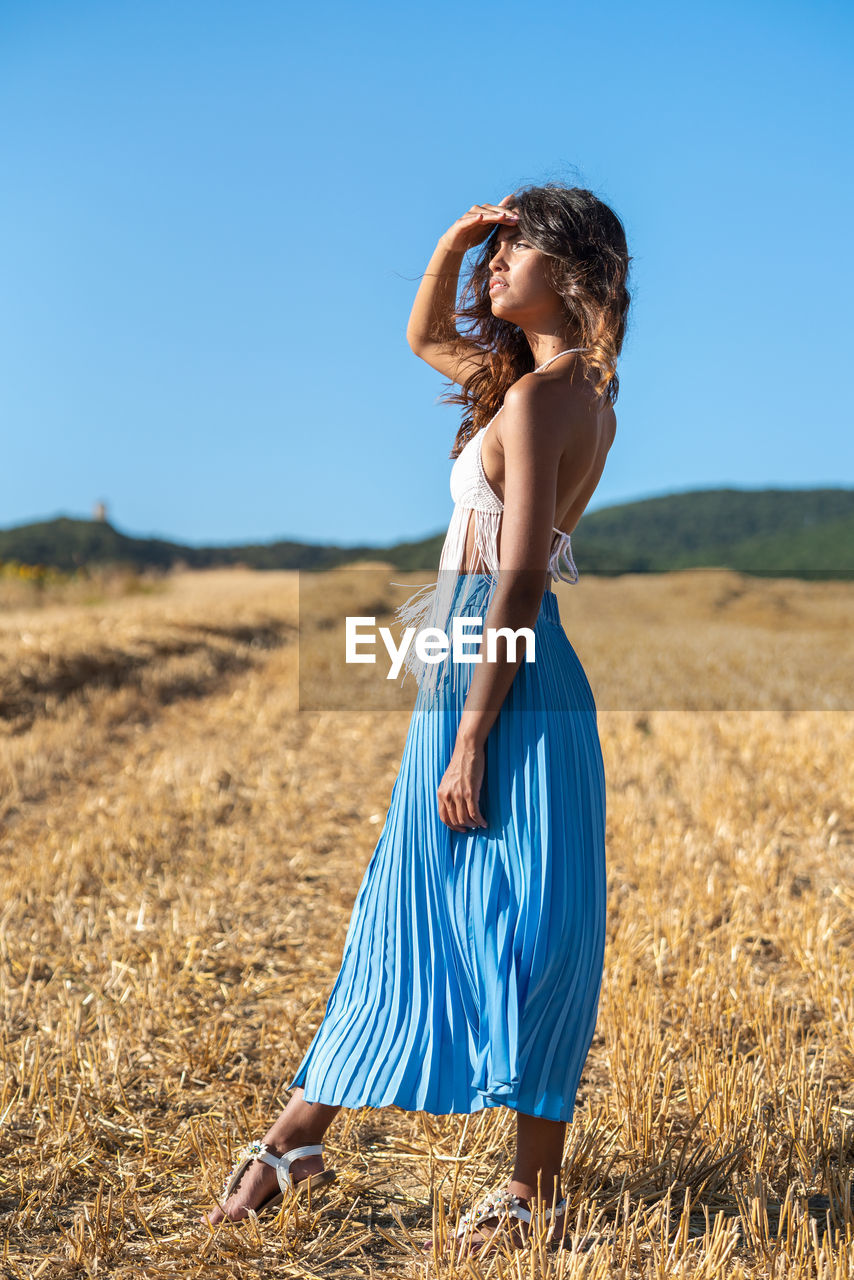 Side view of tranquil female in tassel bra and skirt standing in dry field in summer on sunny day in countryside looking away