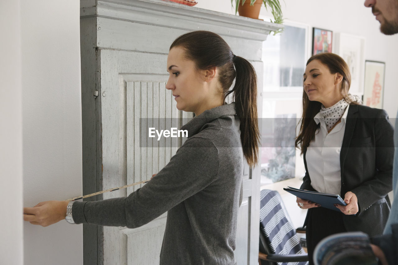 Female realtor and man looking at woman measuring with tape at home