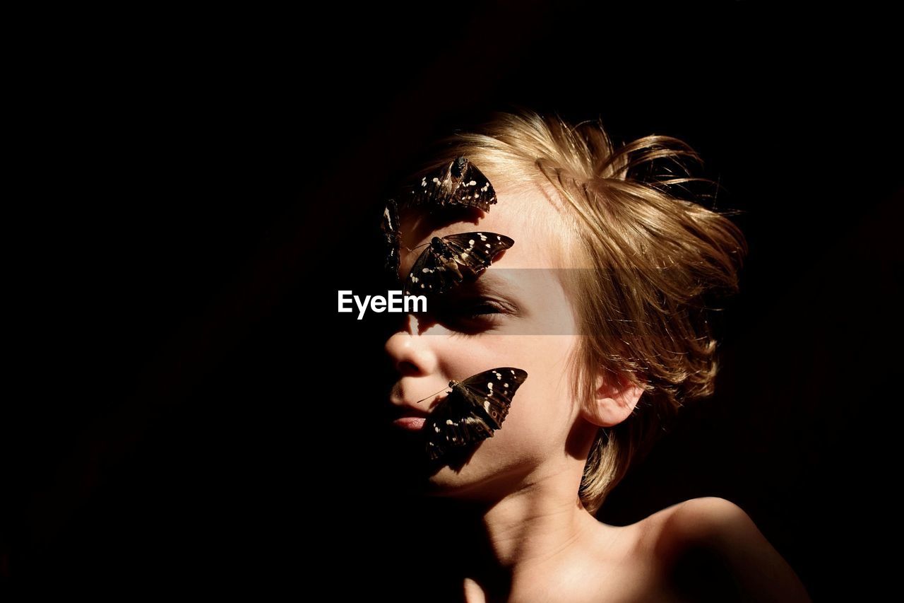 Close-up of boy with butterflies on face against black background