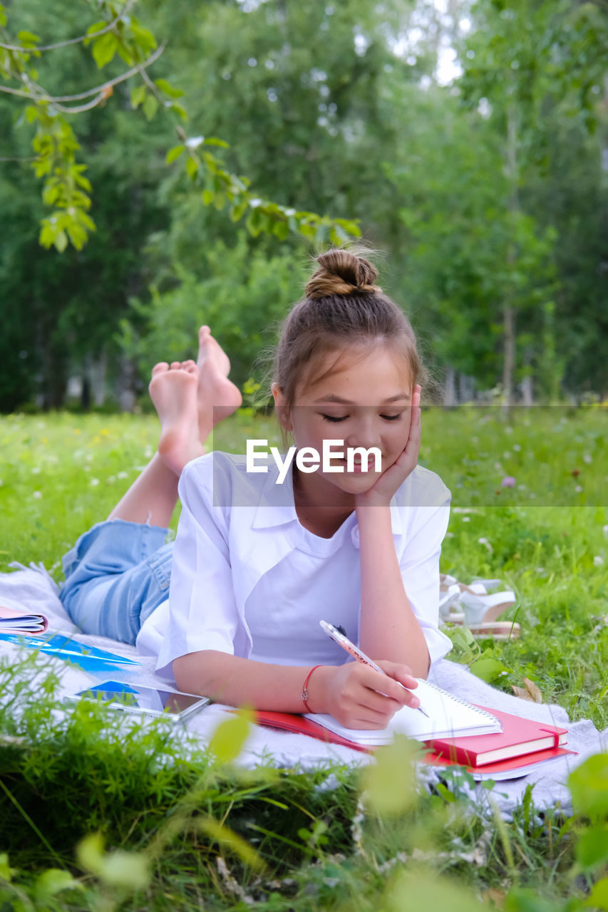 A young schoolgirl lies on the grass in the park with a notebook and a pen.