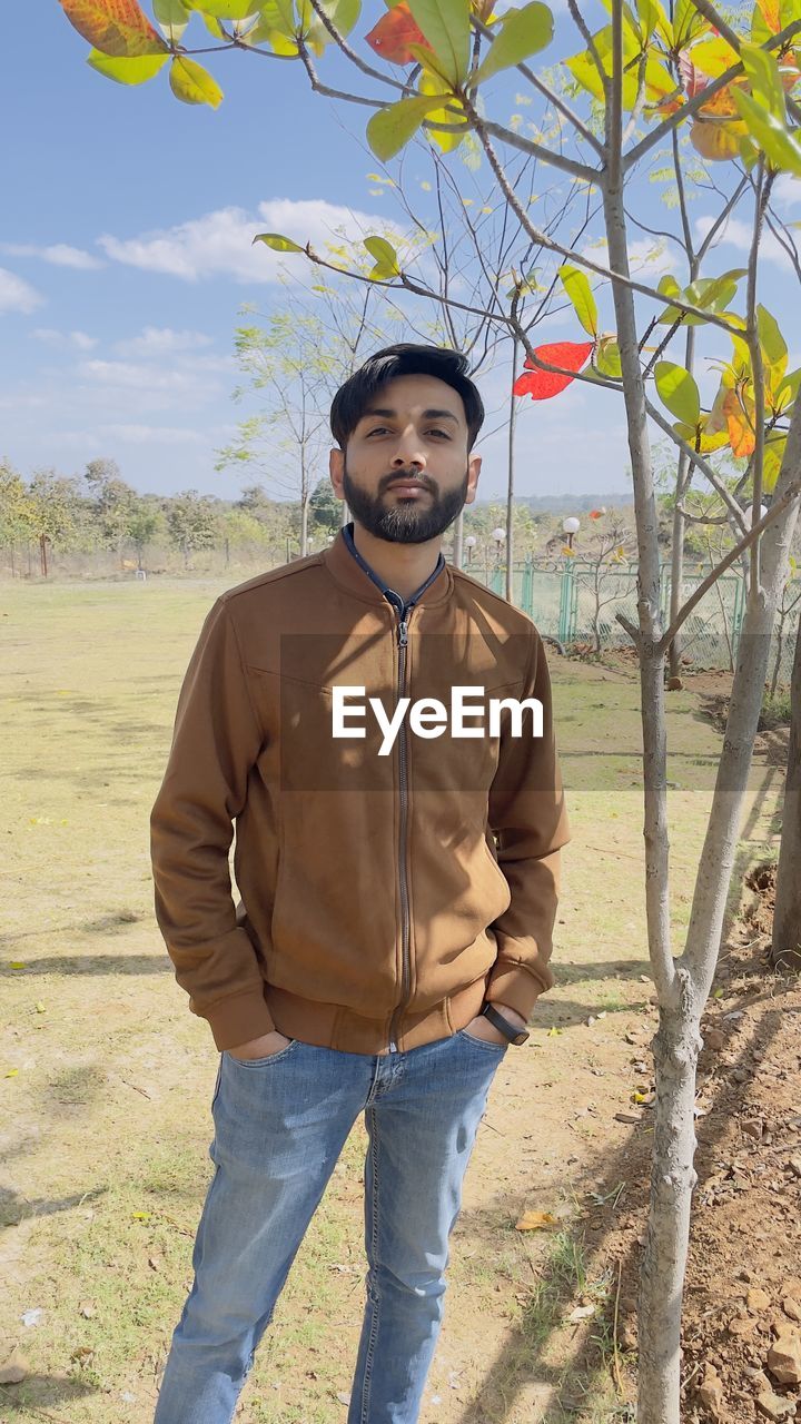 one person, men, standing, nature, adult, portrait, spring, looking at camera, front view, casual clothing, land, plant, young adult, day, clothing, leisure activity, smiling, tree, agriculture, sunlight, landscape, outdoors, lifestyles, hat, sky, facial hair, three quarter length, full length, rural scene, beard, occupation, emotion, happiness, person, jeans, field