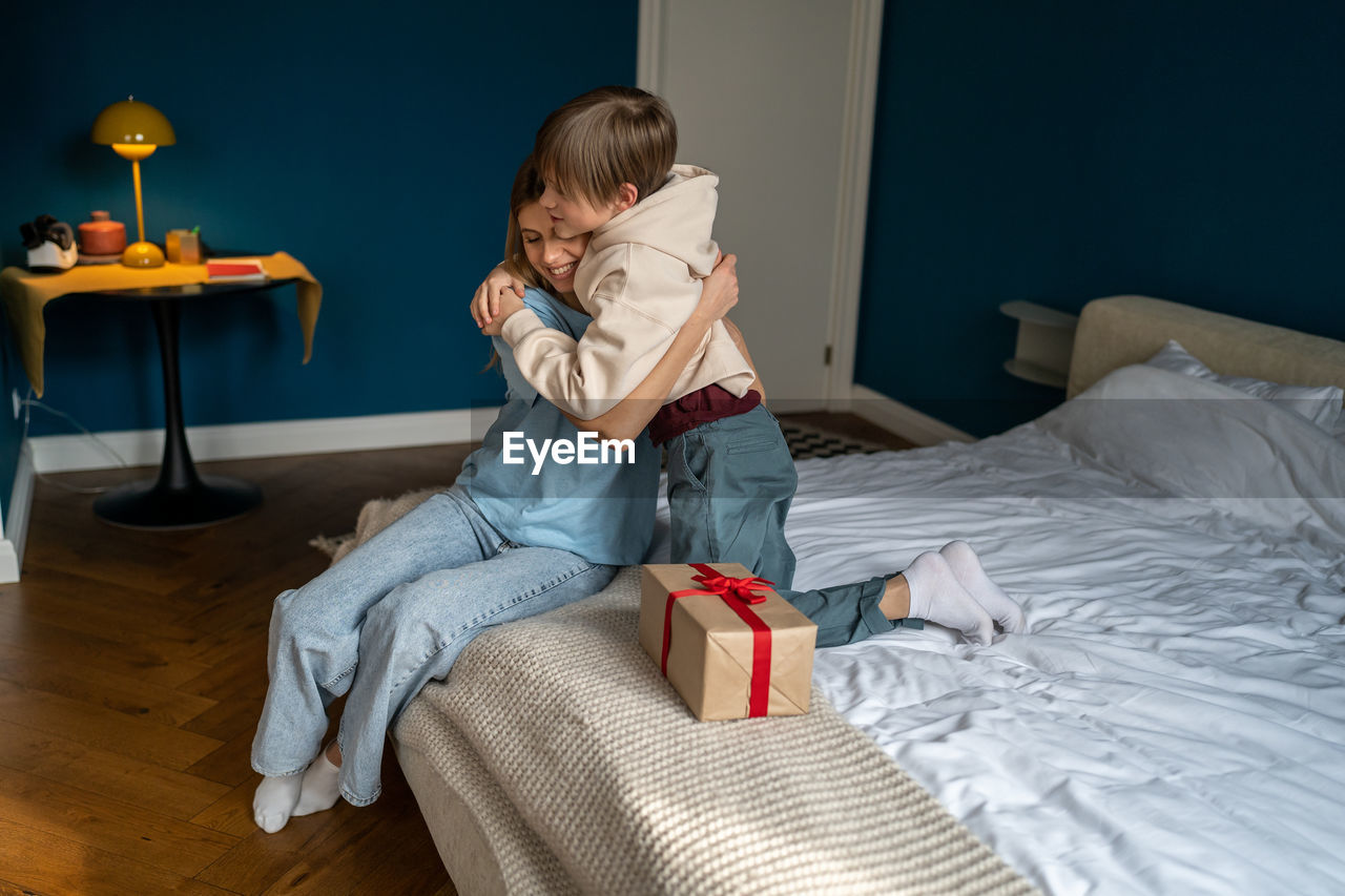Happy little son hugging mother on bed at home, going to unpack festive wrap. birthday concept.