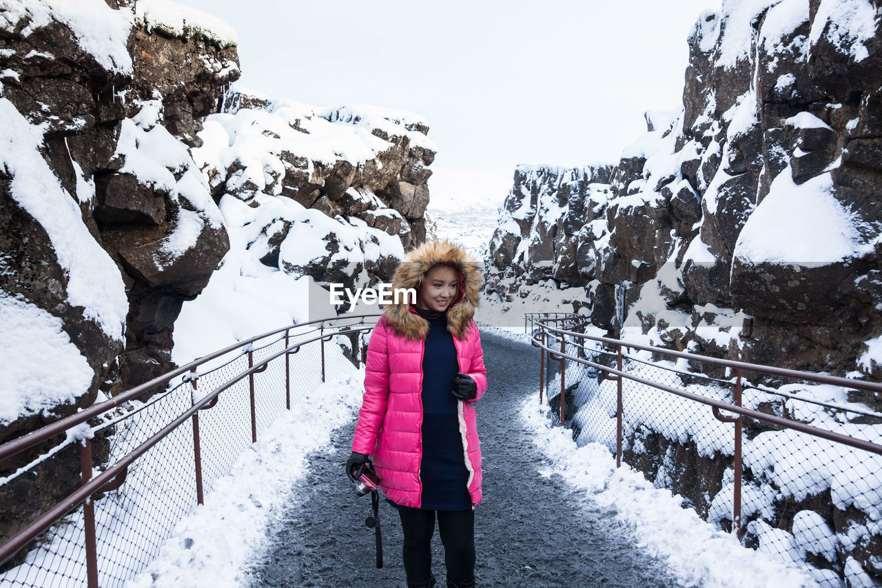 Smiling woman holding camera while standing on road amidst mountain during winter