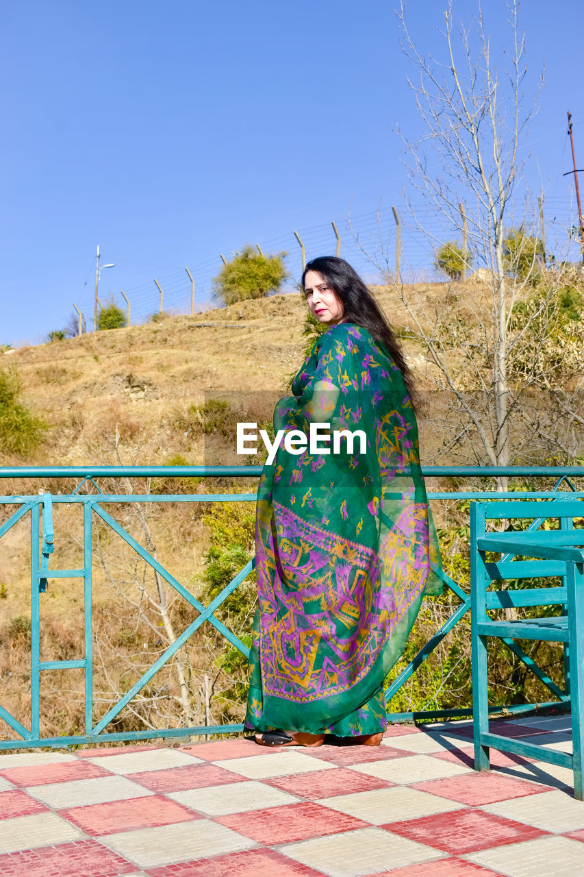 one person, adult, clothing, sky, nature, blue, full length, women, clear sky, fashion, young adult, spring, day, traditional clothing, sunny, plant, outdoors, multi colored, portrait, standing, vacation, lifestyles, pattern, sunlight, looking at camera, front view, leisure activity, person, architecture, land, sitting, female, smiling, landscape