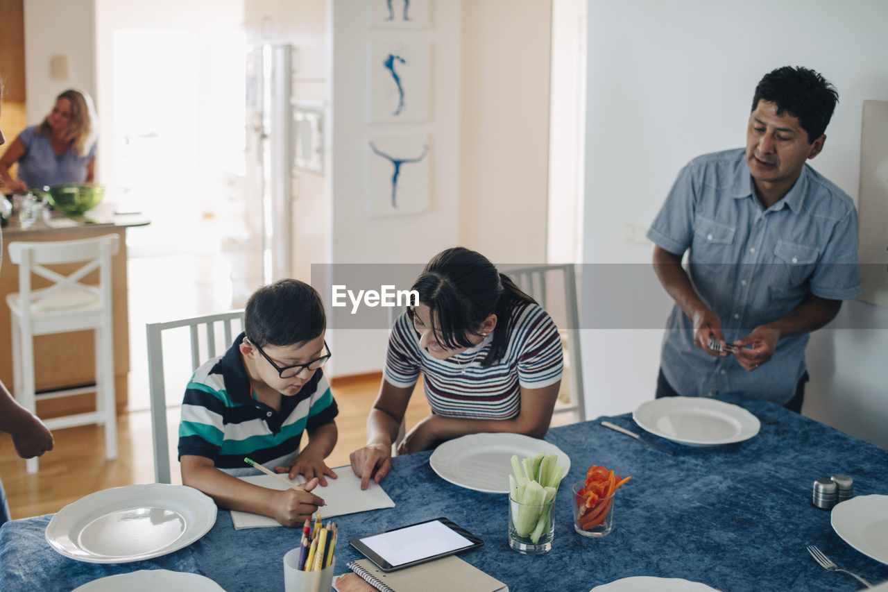 Teenage girl assisting boy in homework while father and sister standing at table