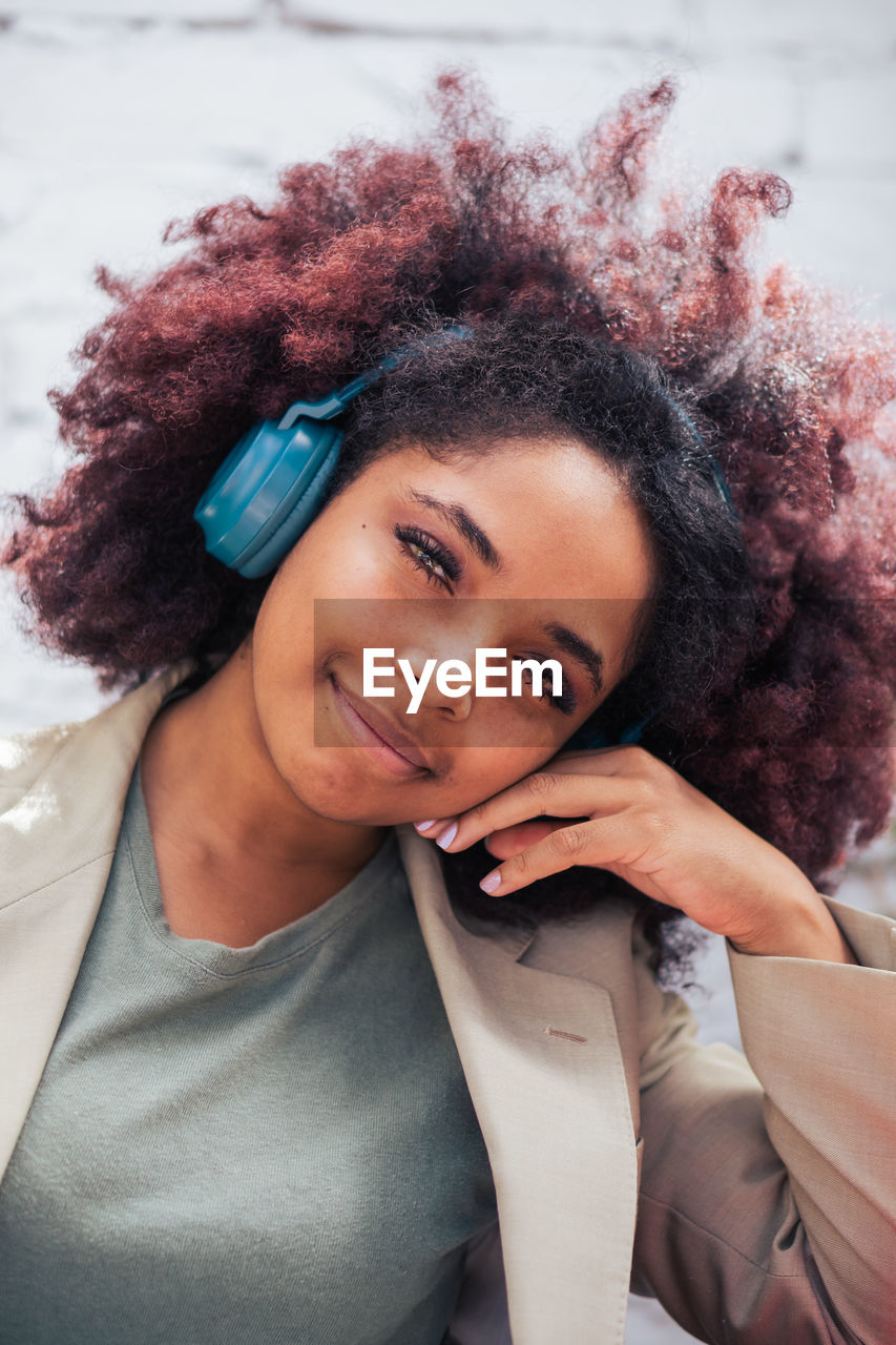 Glad black businesswoman in suit with curly hair touching face and looking at camera while listening to music on street in daytime