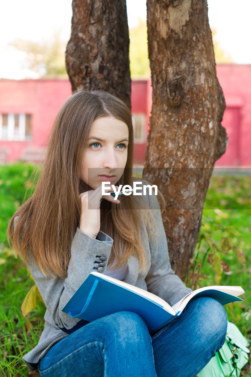 Woman with book and pen sitting against tree in park