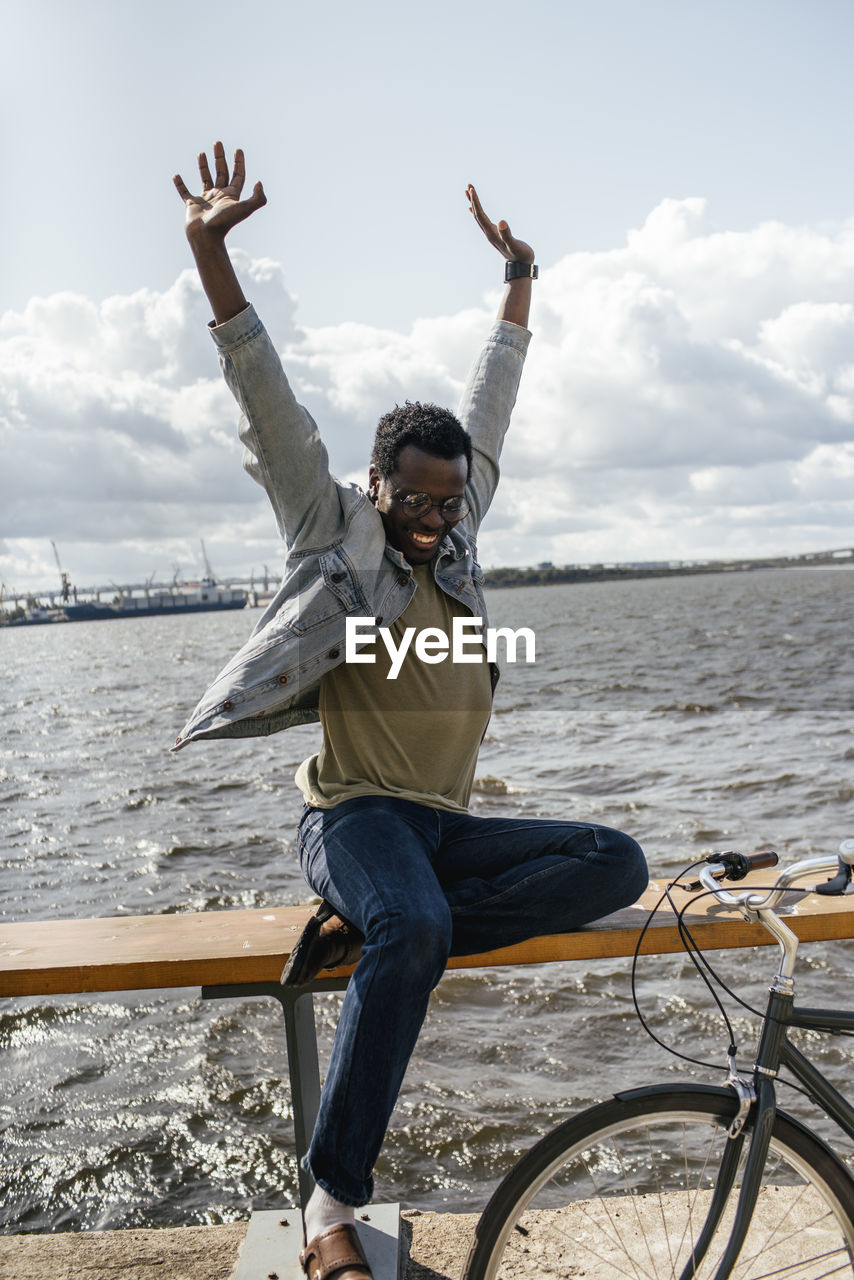 Young man with bicycle, sitting on railng by the sea, pretending to fly