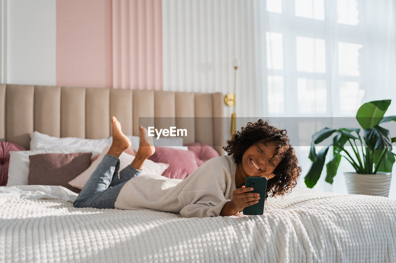 young woman using phone while lying on bed at home