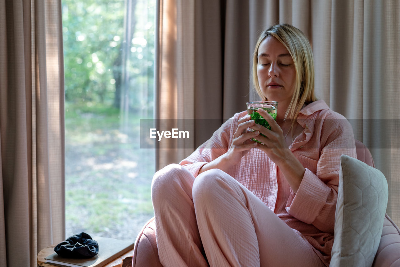 A blond woman in pink pajama sits in the pink armchair and sips mint tea.