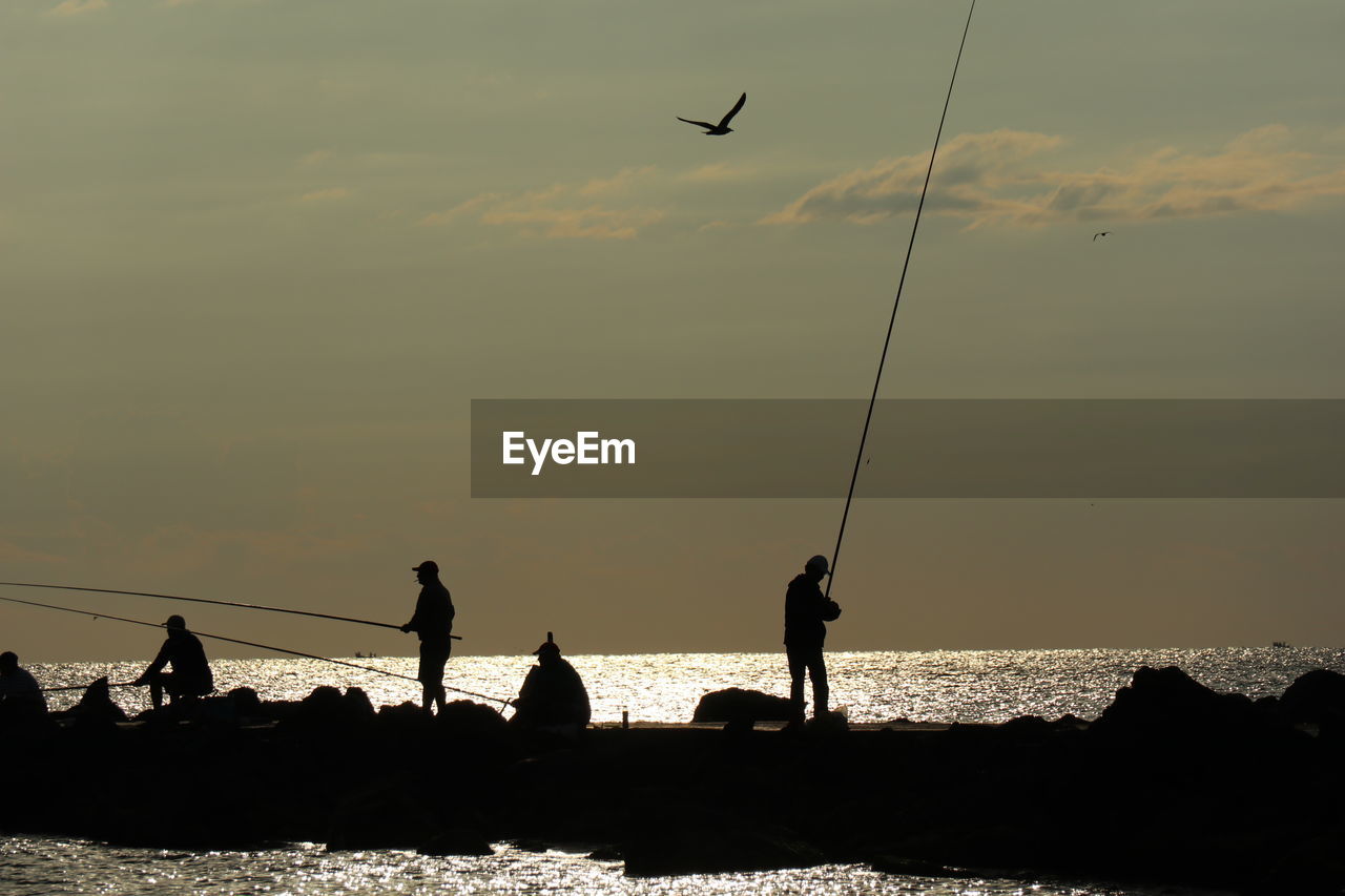 Silhouette people fishing at sea against sky during sunset