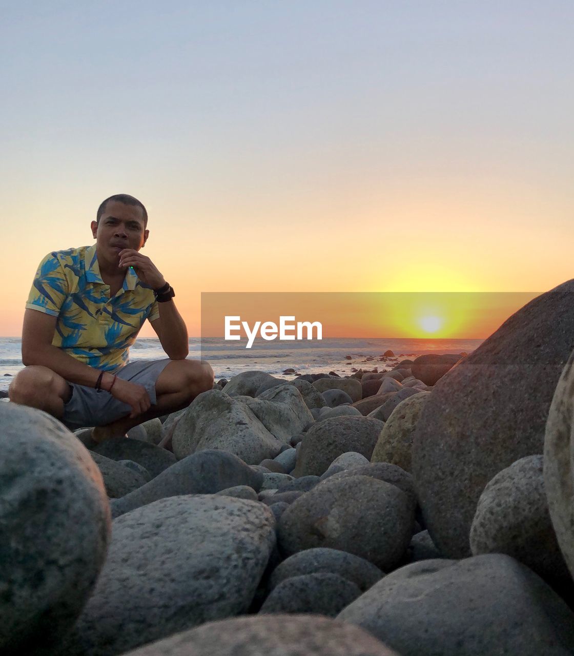 Man crouching on rocks against sea during sunset
