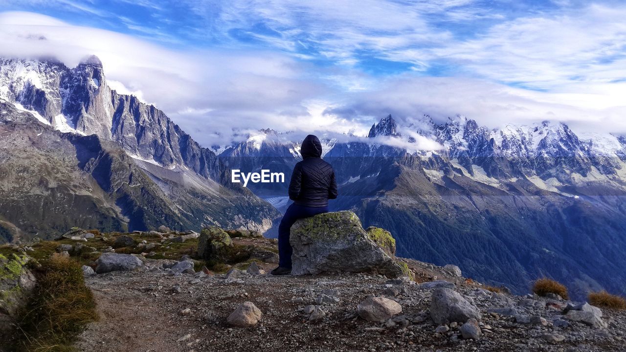 Rear view of woman sitting on rock while looking at mountains against sky