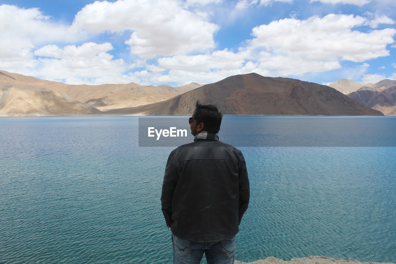 Rear view of man looking at mountains against sky while standing by lake