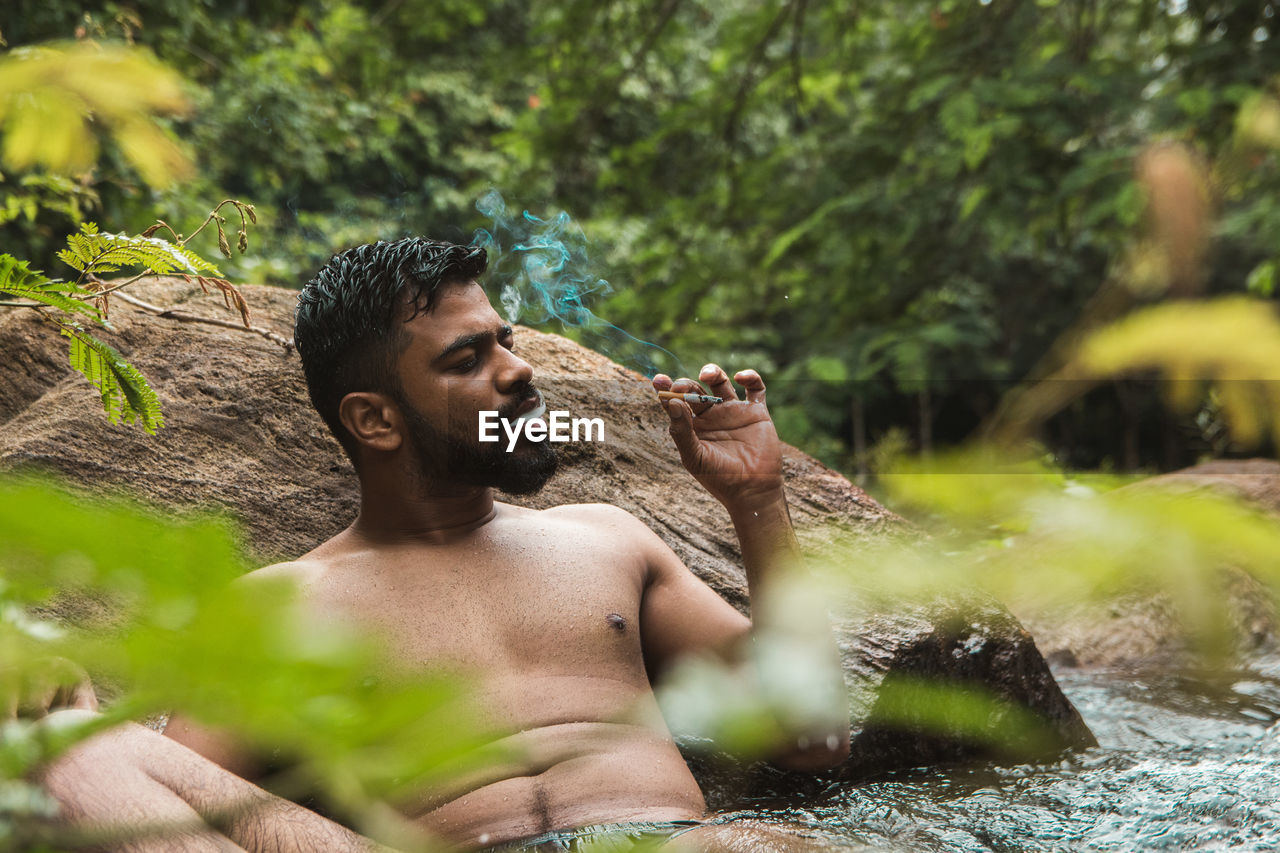 Shirtless man smoking while relaxing by rock in river