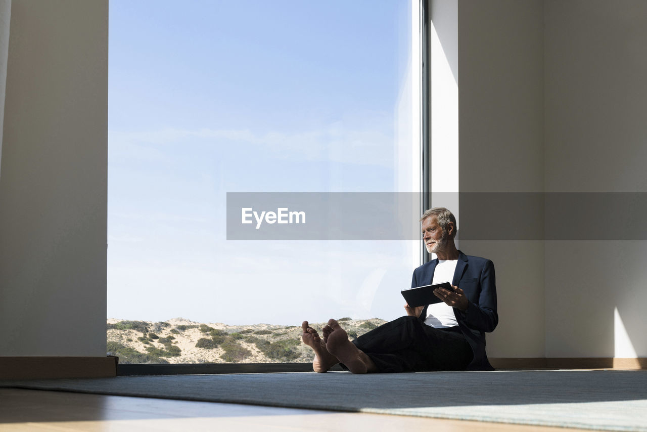 Senior businessman sitting at panorama window using tablet