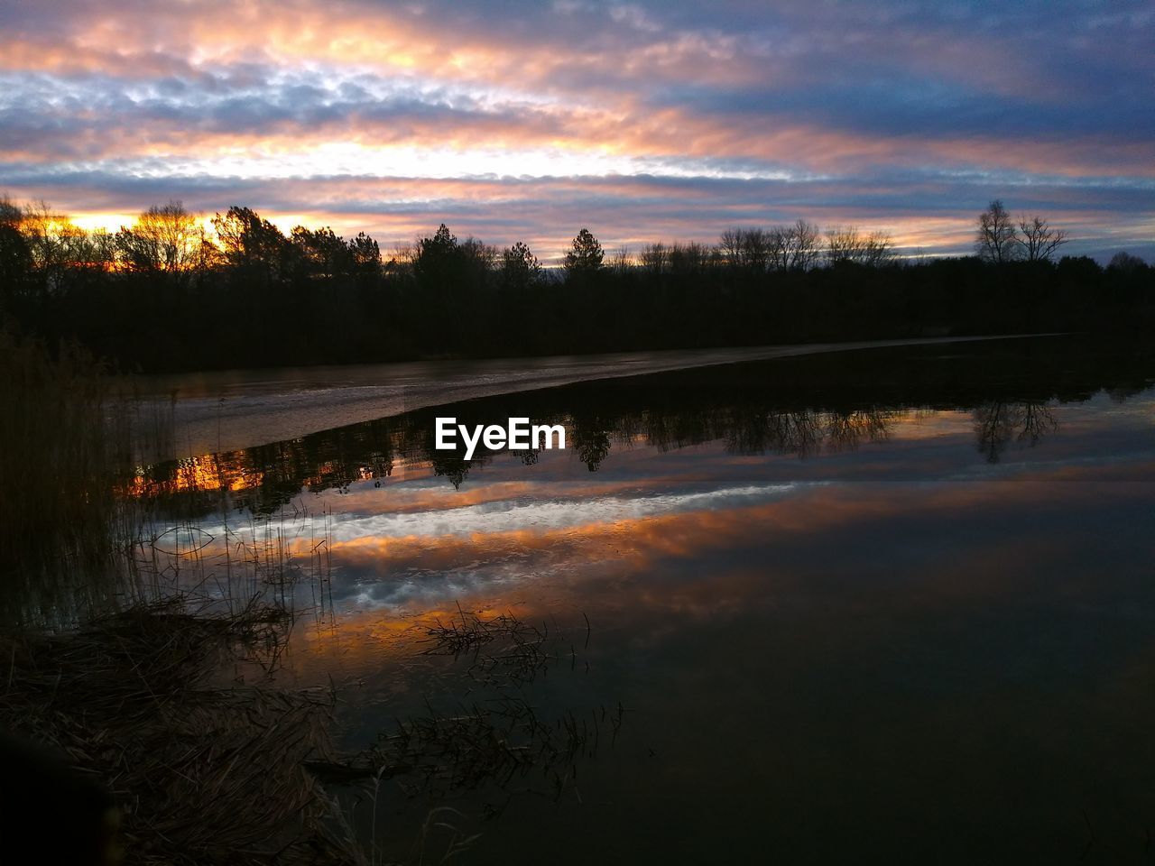 REFLECTION OF SILHOUETTE TREES IN LAKE AGAINST SKY DURING SUNSET