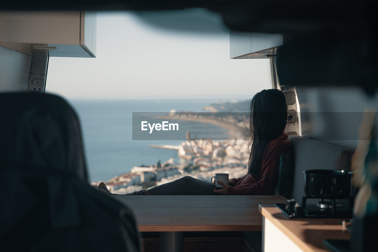 Rear view of woman sitting at table in bus by sea against sky