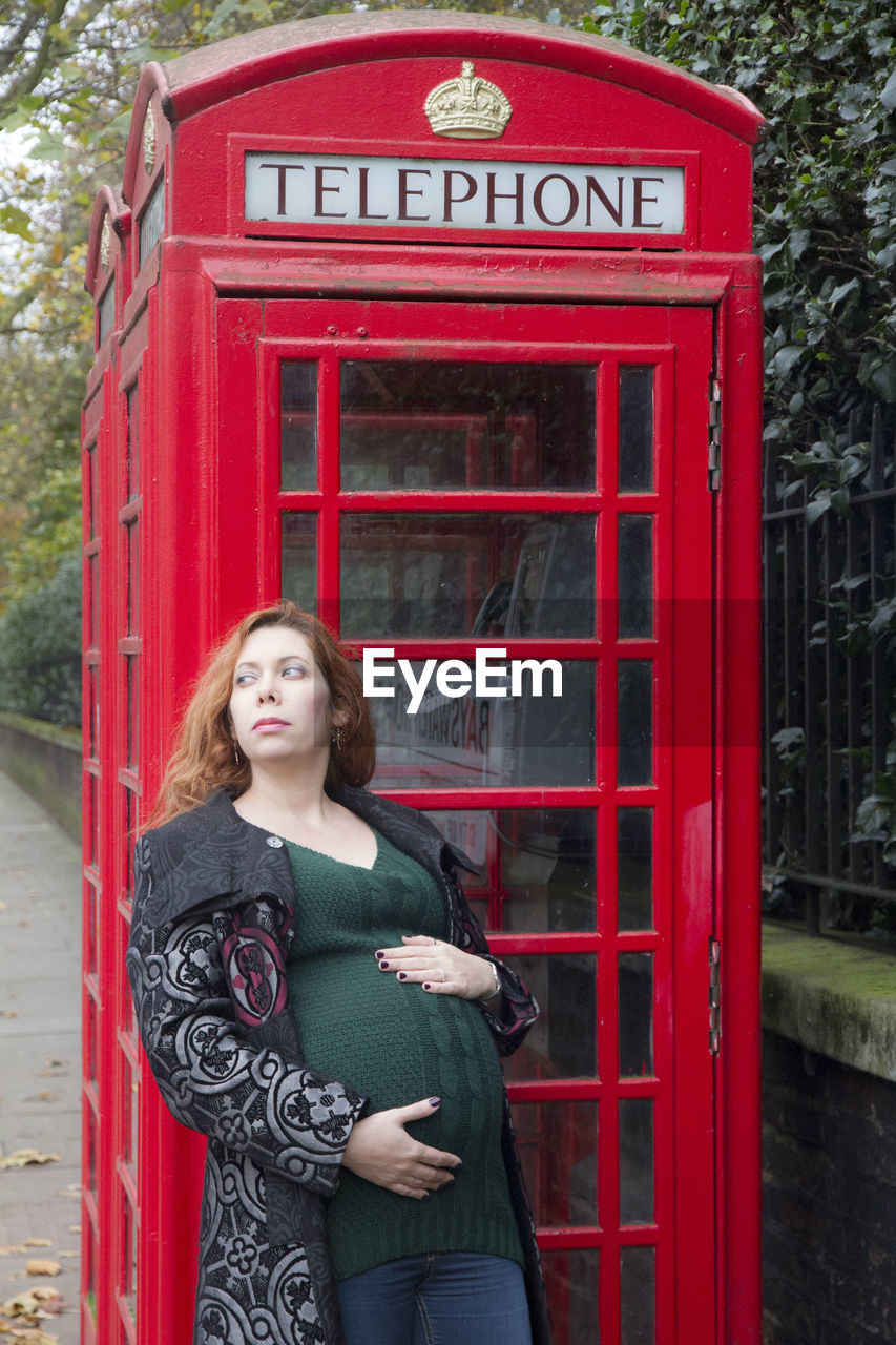 Thoughtful pregnant woman leaning on telephone booth at sidewalk