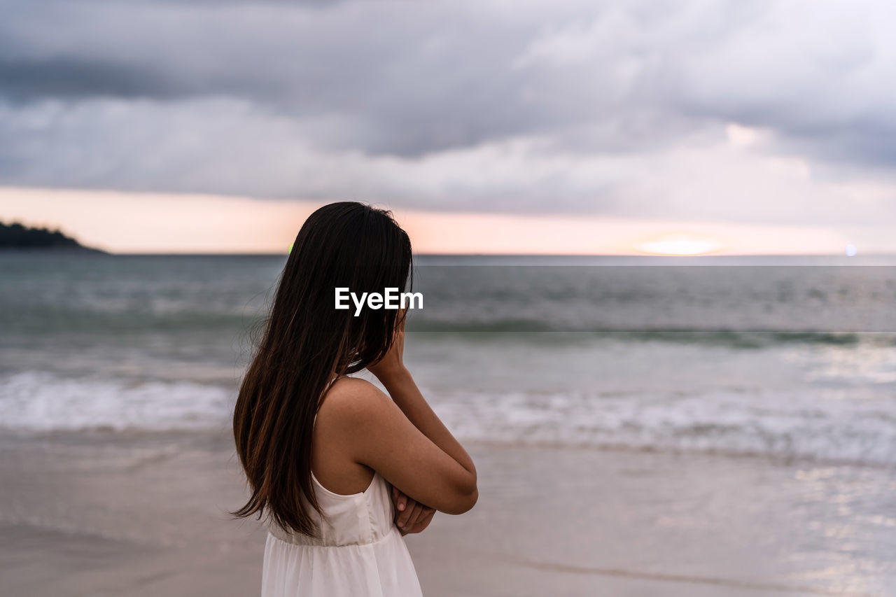 side view of young woman standing at beach against sky during sunset