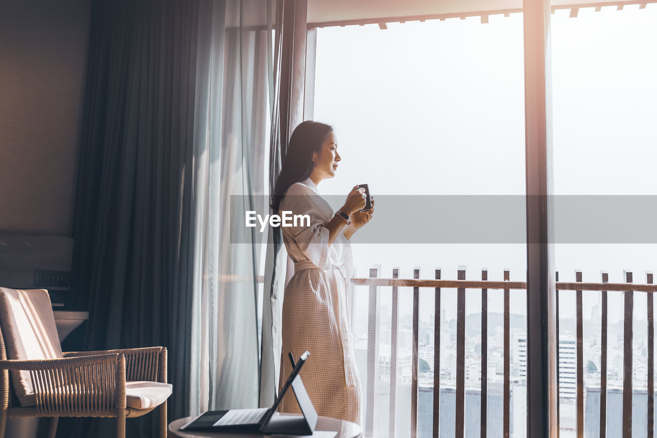 Side view of woman looking through window at hotel room