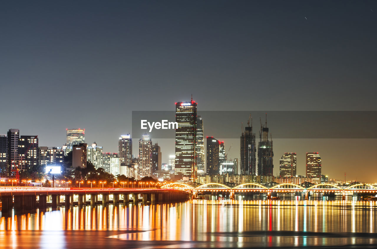 Illuminated buildings by river against sky at night