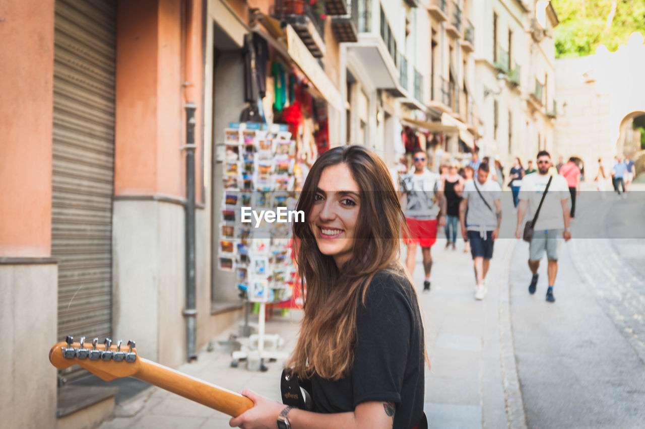 Portrait of young woman playing guitar while standing on sidewalk in city