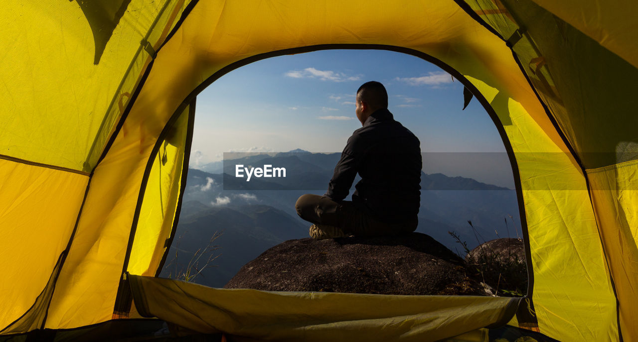 Rear view of man sitting on mountain by tent against sky